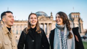Three students stand smiling on the lawn in front of the Reichstag in Berlin.